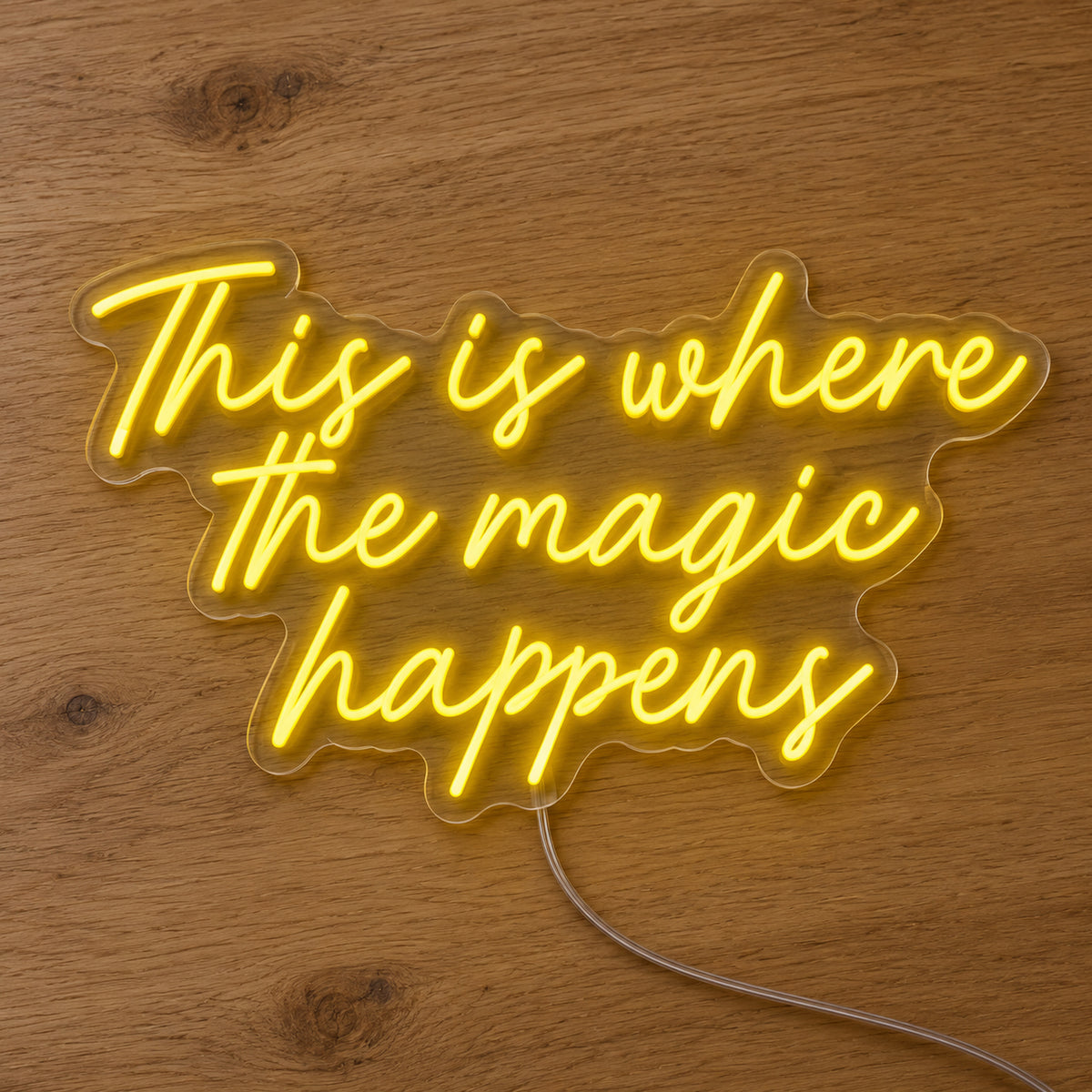 This Is Where The Magic Happens Neon Sign in yellow, featuring three lines of script lettering on a wooden surface.