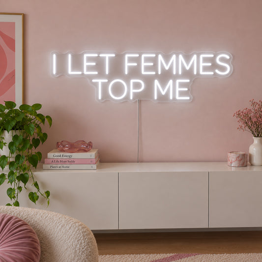 I Let Femmes Top Me Neon Sign glowing white in rounded lettering above a white sideboard.
