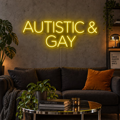 Autistic & Gay Neon Sign with yellow lighting, displayed above a dark sofa against a concrete wall.
