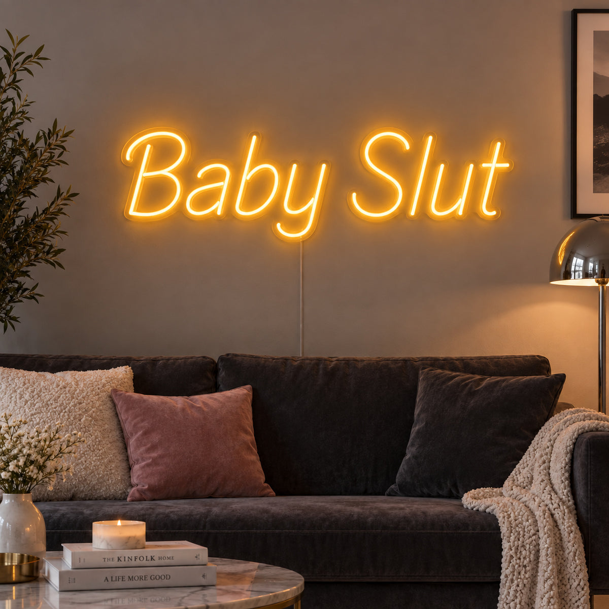 Baby Slut Neon Sign glowing gold above a grey sofa with pink cushions.
