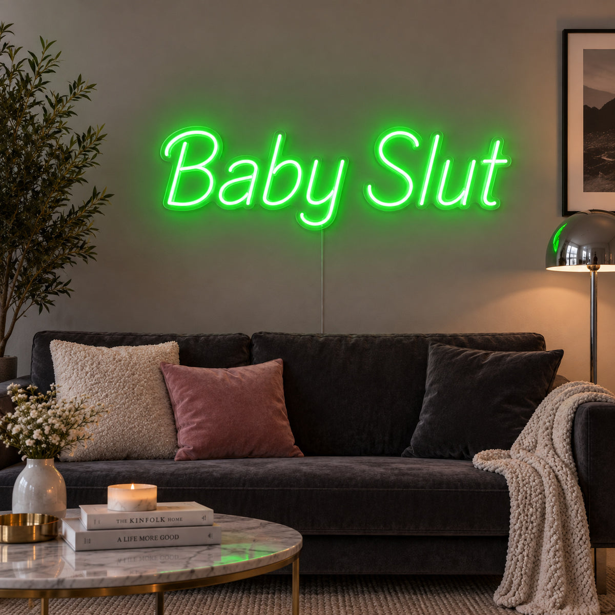 Baby Slut Neon Sign with green lighting, displayed above a grey sofa with pink cushions.