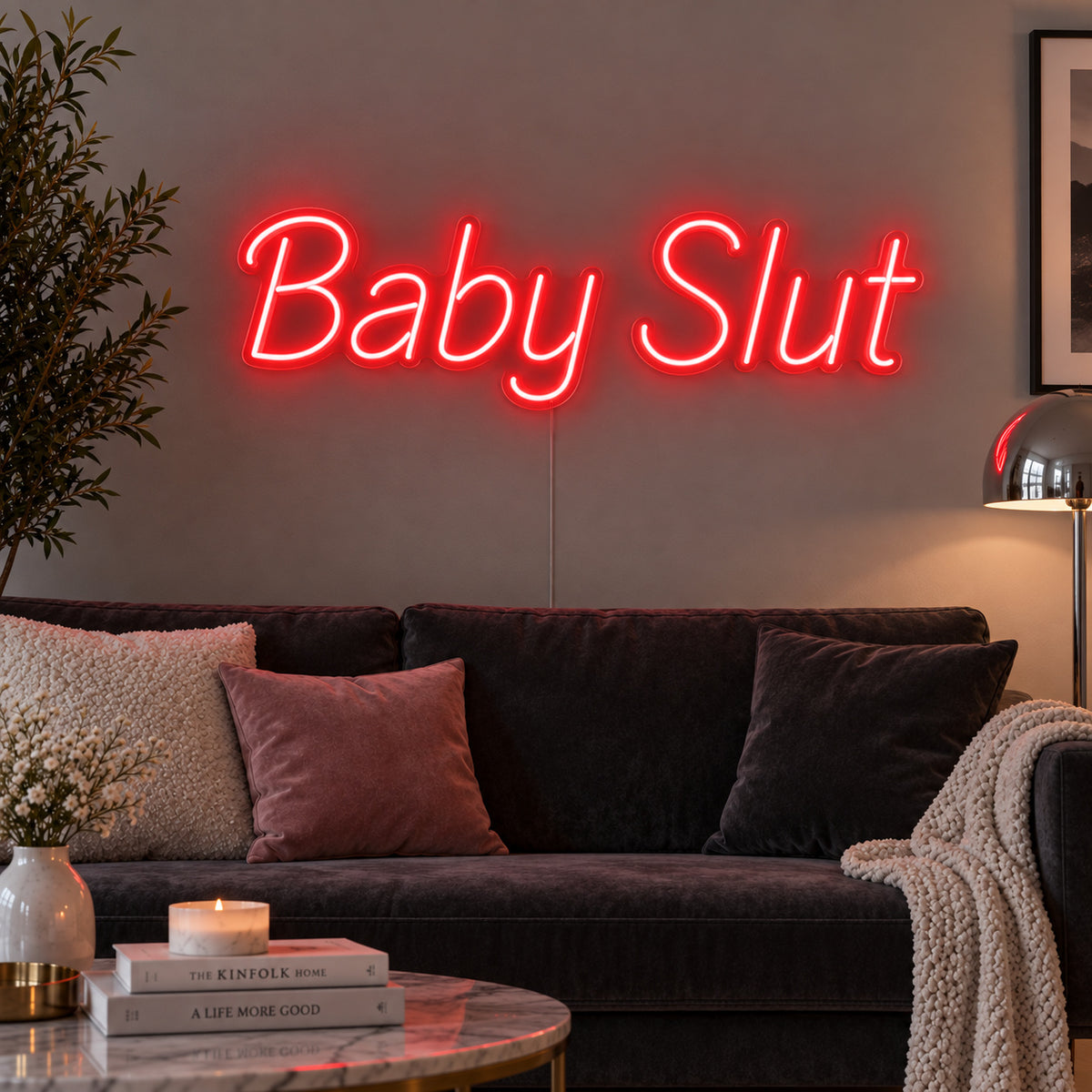 Baby Slut Neon Sign with red lighting, displayed above a grey sofa with pink cushions.