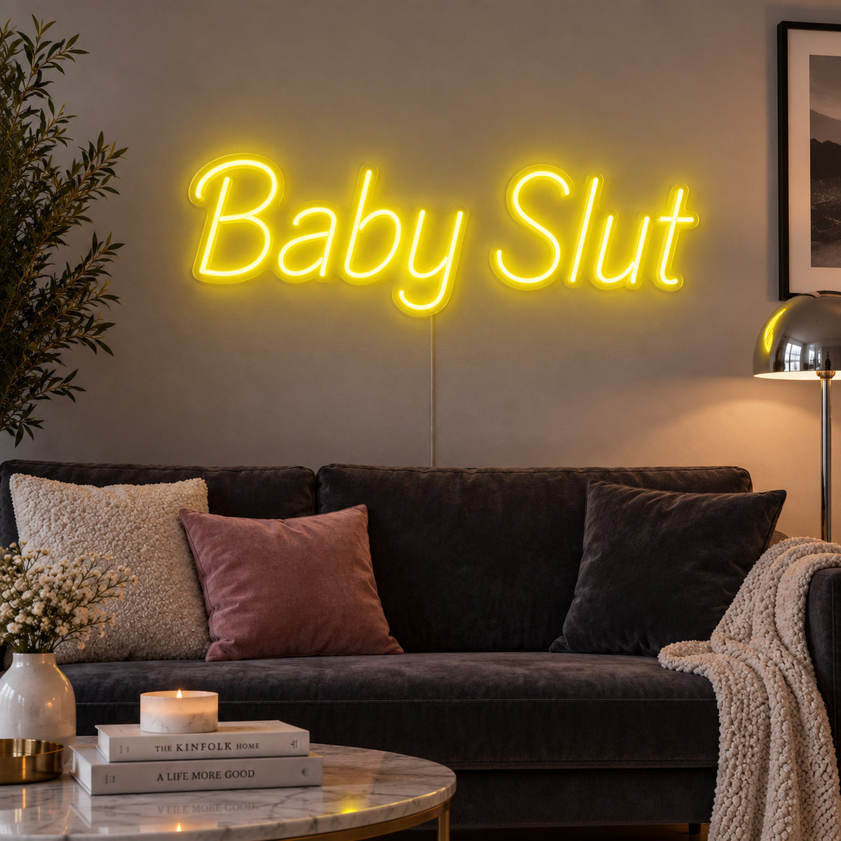 Baby Slut Neon Sign with yellow lighting, displayed above a grey sofa with pink cushions.