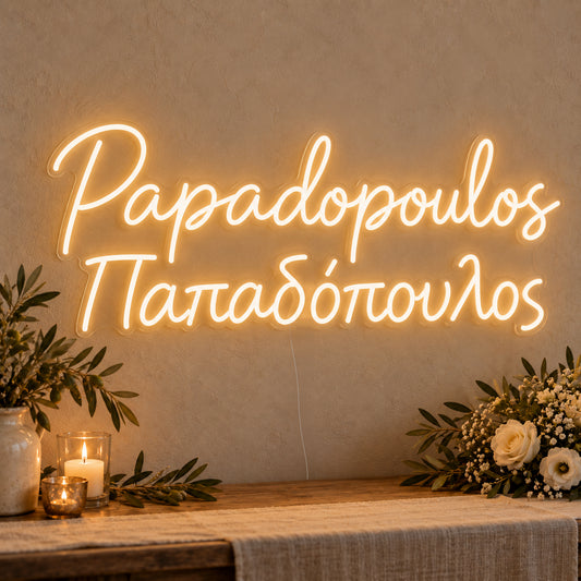 Personalised Bilingual Greek Surname Neon Sign on clear cut-to-shape acrylic in an illuminated wall display; example personalisation shown.
