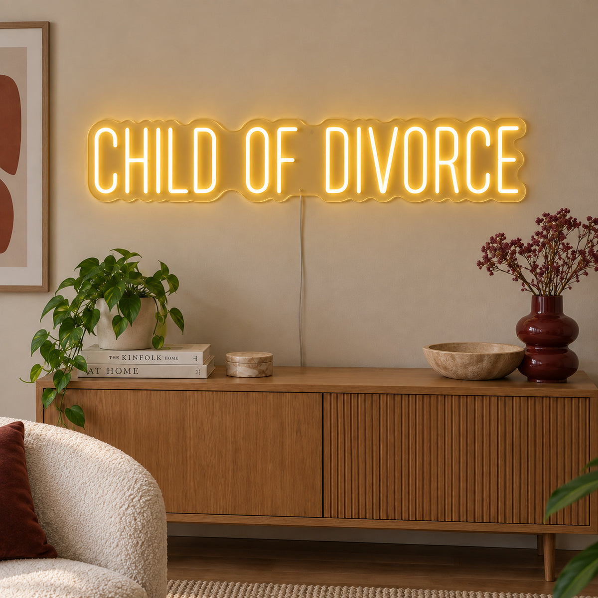 Child Of Divorce Neon Sign glowing gold above a wooden sideboard with plants.