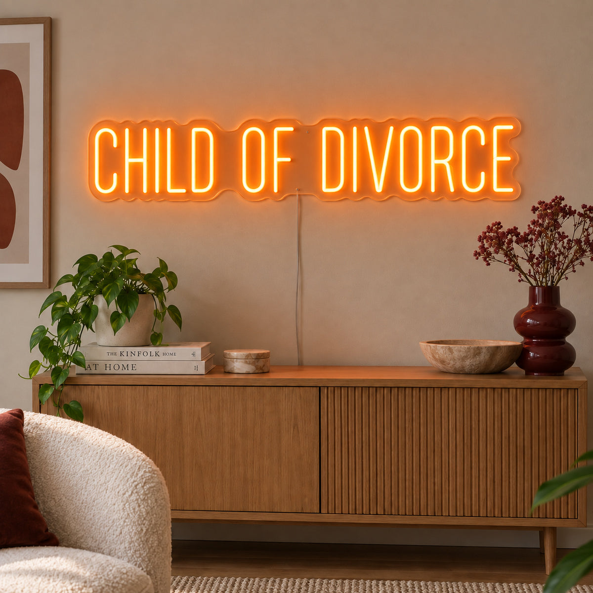 Orange version of the Child Of Divorce Neon Sign, shown above a wooden sideboard with plants.