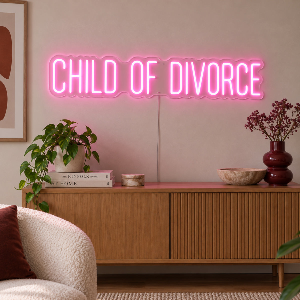 Child Of Divorce Neon Sign glowing pink above a wooden sideboard with plants.
