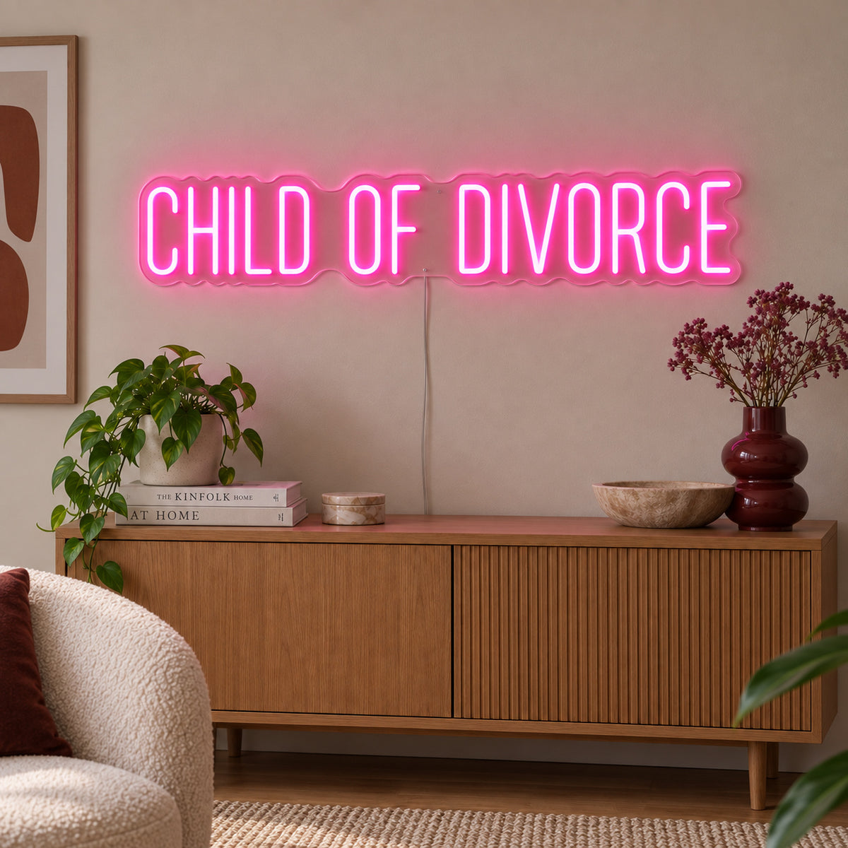Rose Red version of the Child Of Divorce Neon Sign, shown above a wooden sideboard with plants.