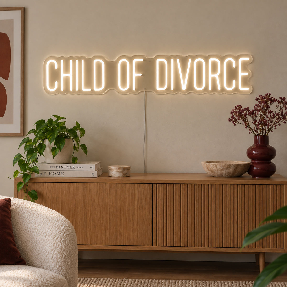 Warm White version of the Child Of Divorce Neon Sign, shown above a wooden sideboard with plants.