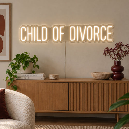 Warm White version of the Child Of Divorce Neon Sign, shown above a wooden sideboard with plants.