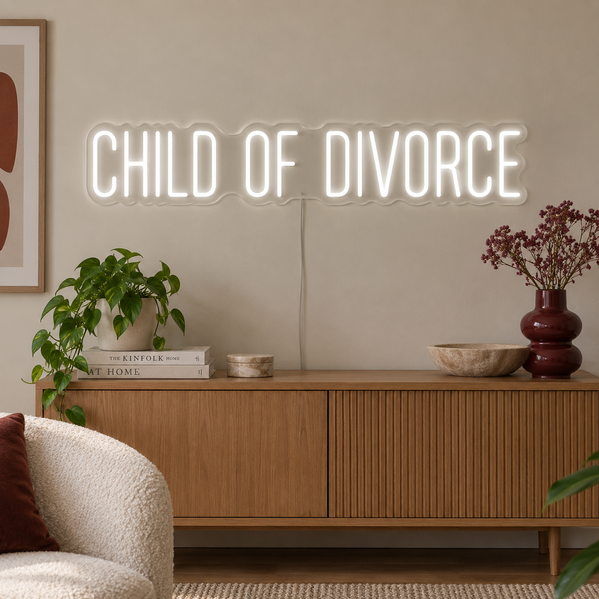 Child Of Divorce Neon Sign in white above a wooden sideboard with plants.