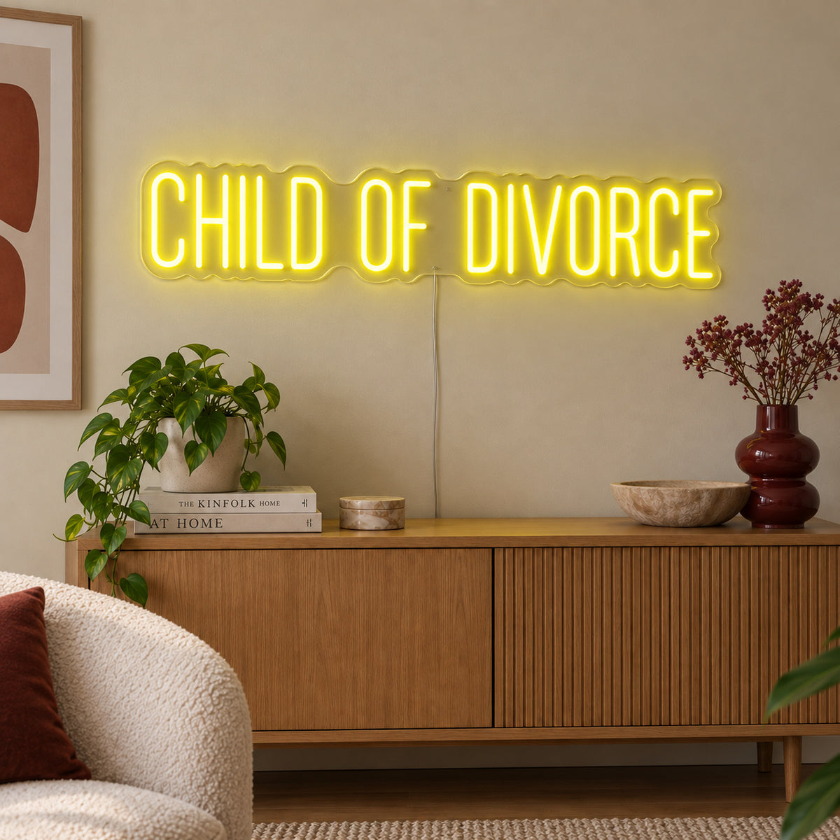 Child Of Divorce Neon Sign with yellow lighting, displayed above a wooden sideboard with plants.