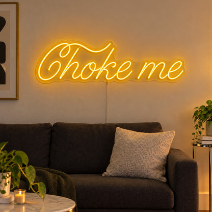 Choke Me Neon Sign in yellow, featuring Choke Me script lettering above a dark sofa.