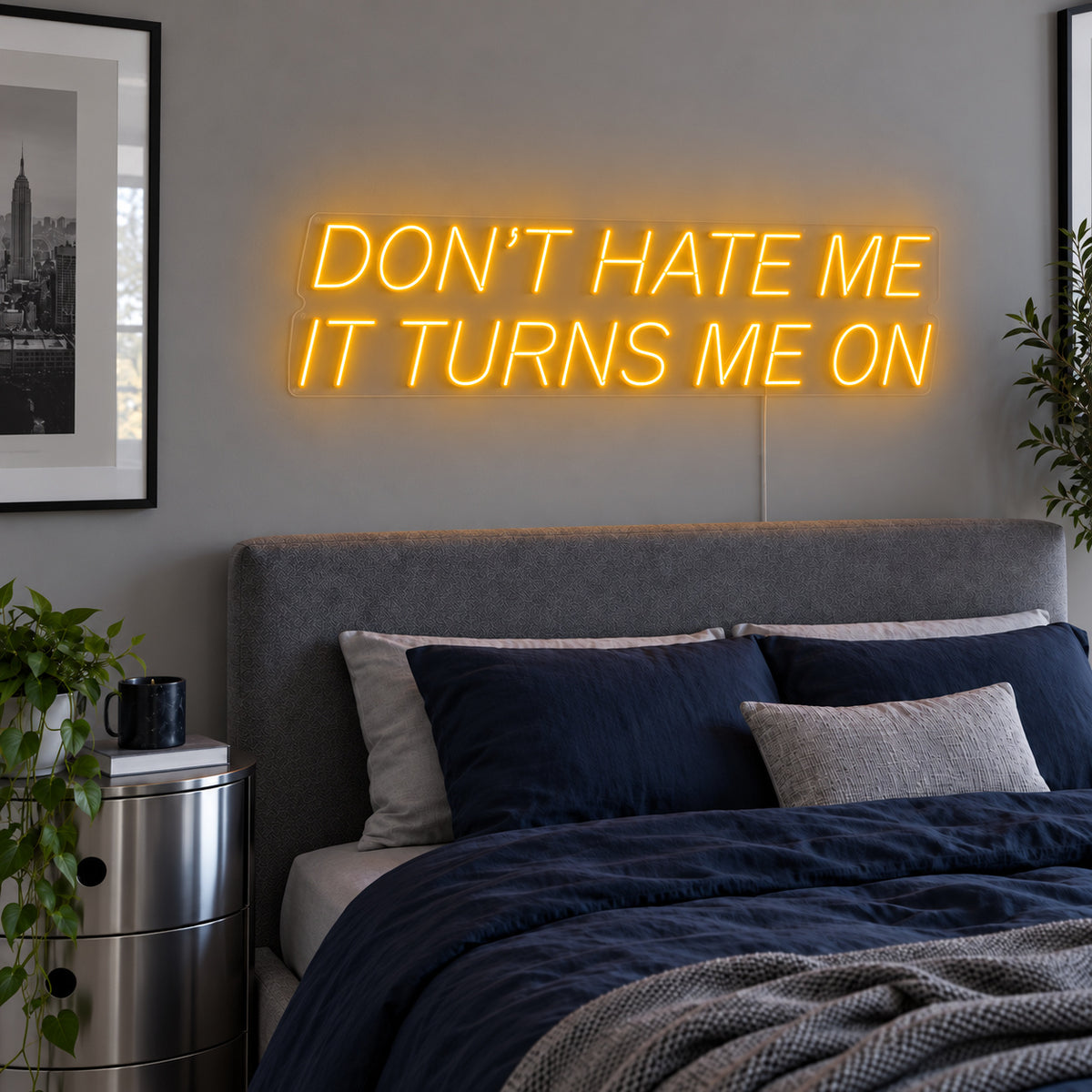 Don't Hate Me It Turns Me On Neon Sign glowing gold above a bed with dark blue bedding.