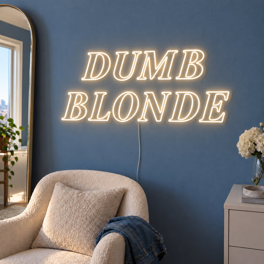 Warm White version of the Dumb Blonde Neon Sign, shown in outlined serif lettering above a cream chair.