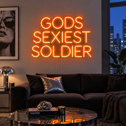 Orange version of the Gods Sexiest Soldier Neon Sign, shown in rounded lettering above a dark sofa.