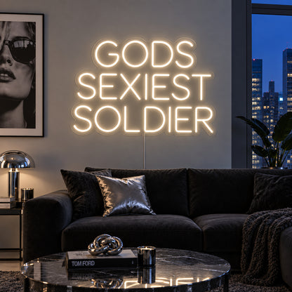 Warm White version of the Gods Sexiest Soldier Neon Sign, shown in rounded lettering above a dark sofa.