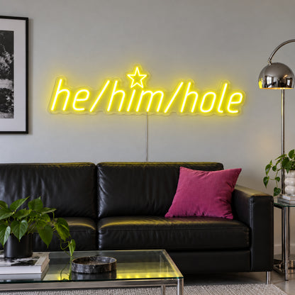 He/Him/Hole Neon Sign with yellow lighting, displayed above a black leather sofa.