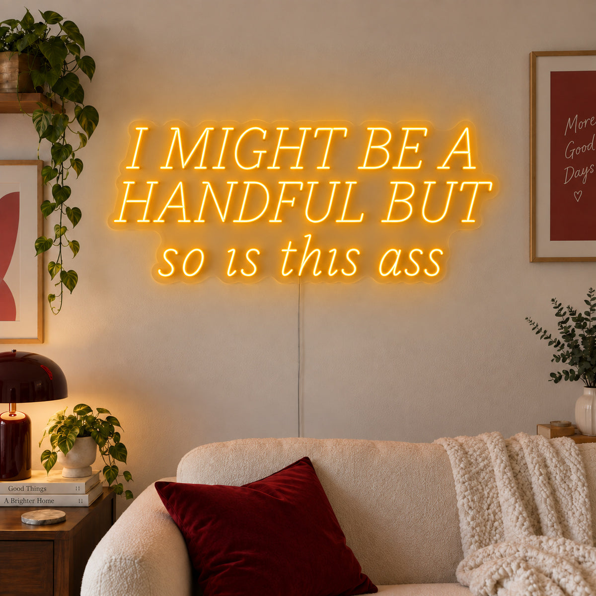 I Might Be A Handful But So Is This Ass Neon Sign glowing gold in wide lettering above a cream sofa.