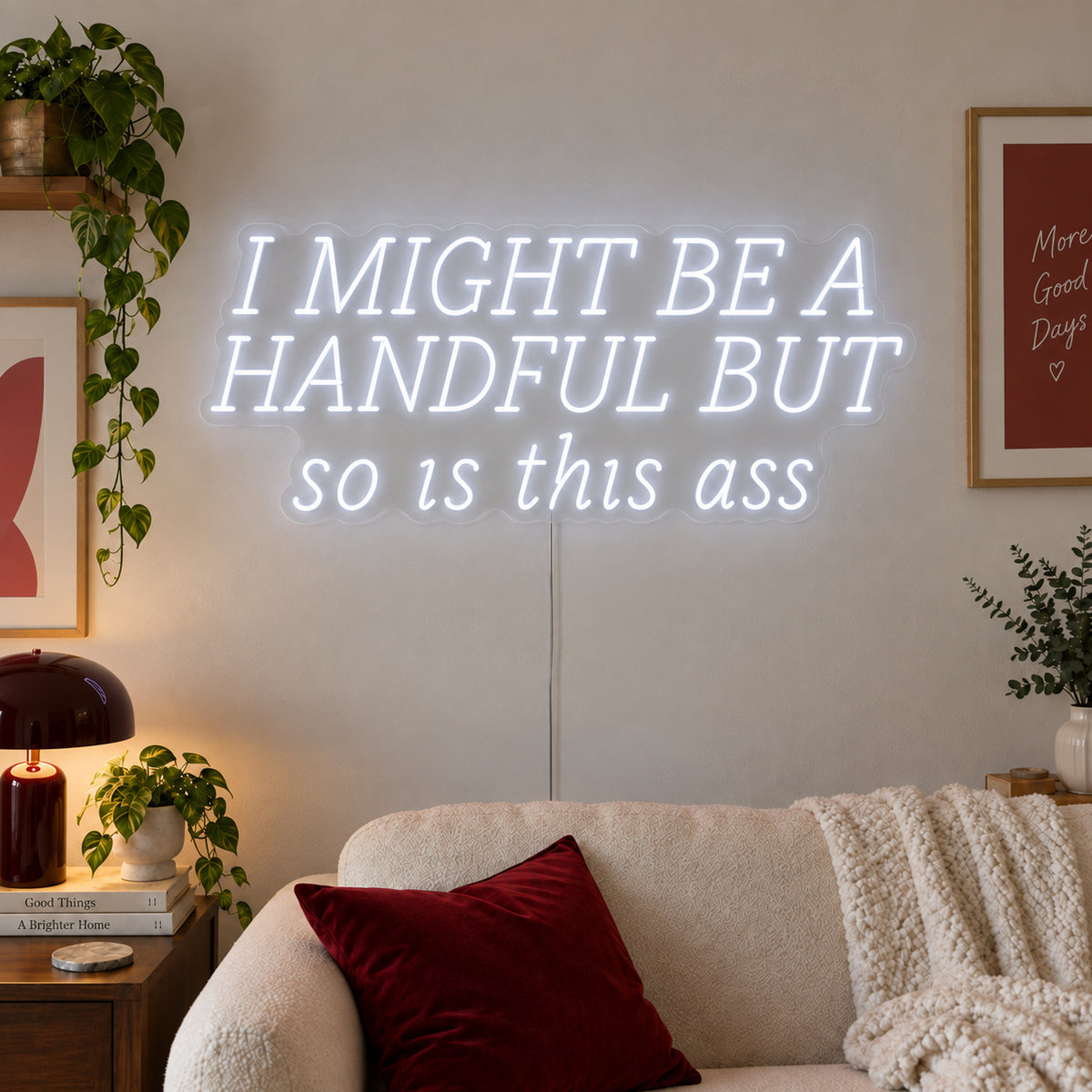 I Might Be A Handful But So Is This Ass Neon Sign glowing white in wide lettering above a cream sofa.