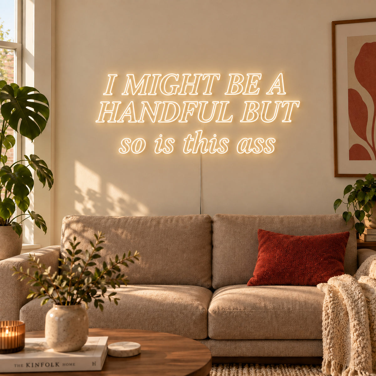 Warm White version of the I Might Be A Handful But So Is This Ass Neon Sign, shown in compact lettering above a cream sofa.
