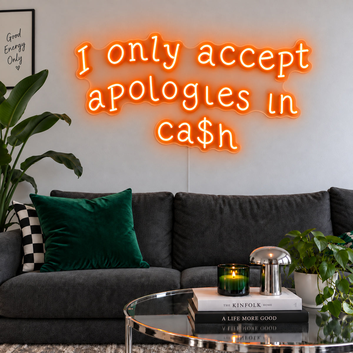 Orange version of the I Only Accept Apologies In Ca$h Neon Sign, shown above a charcoal sofa with green cushions.