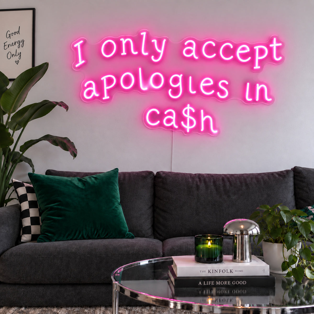 I Only Accept Apologies In Ca$h Neon Sign glowing pink above a charcoal sofa with green cushions.