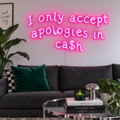 I Only Accept Apologies In Ca$h Neon Sign glowing pink above a charcoal sofa with green cushions.