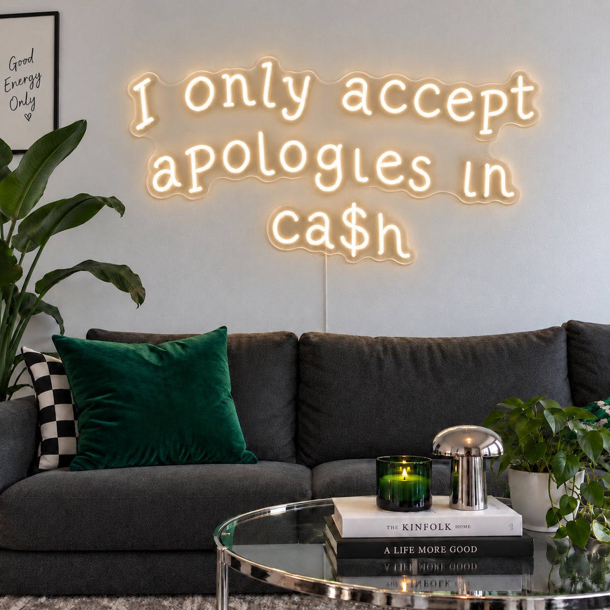 Warm White version of the I Only Accept Apologies In Ca$h Neon Sign, shown above a charcoal sofa with green cushions.