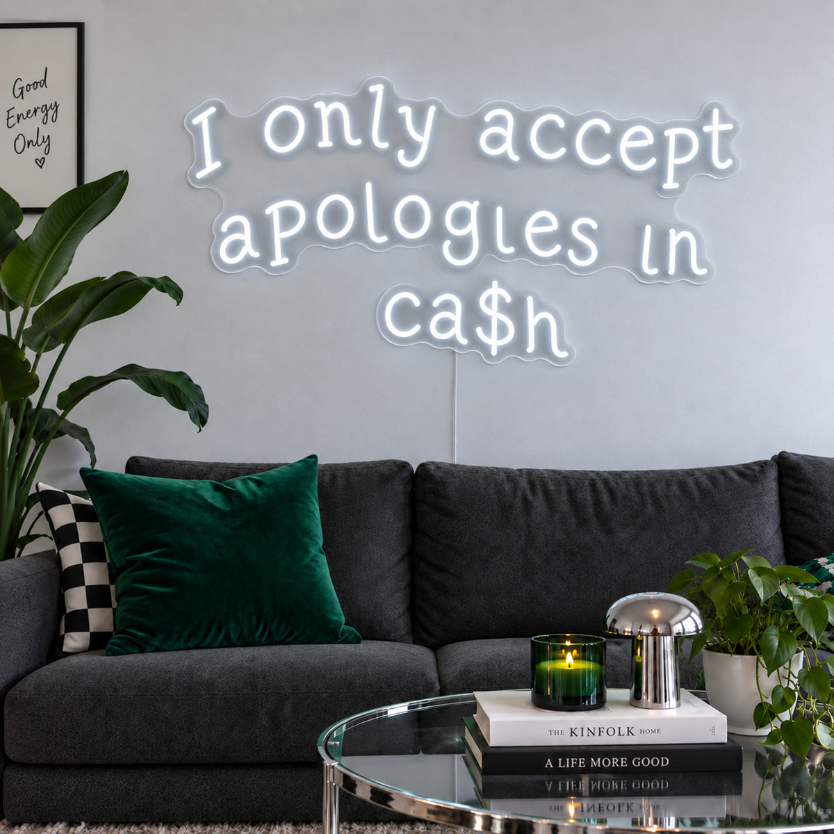 I Only Accept Apologies In Ca$h Neon Sign glowing white above a charcoal sofa with green cushions.
