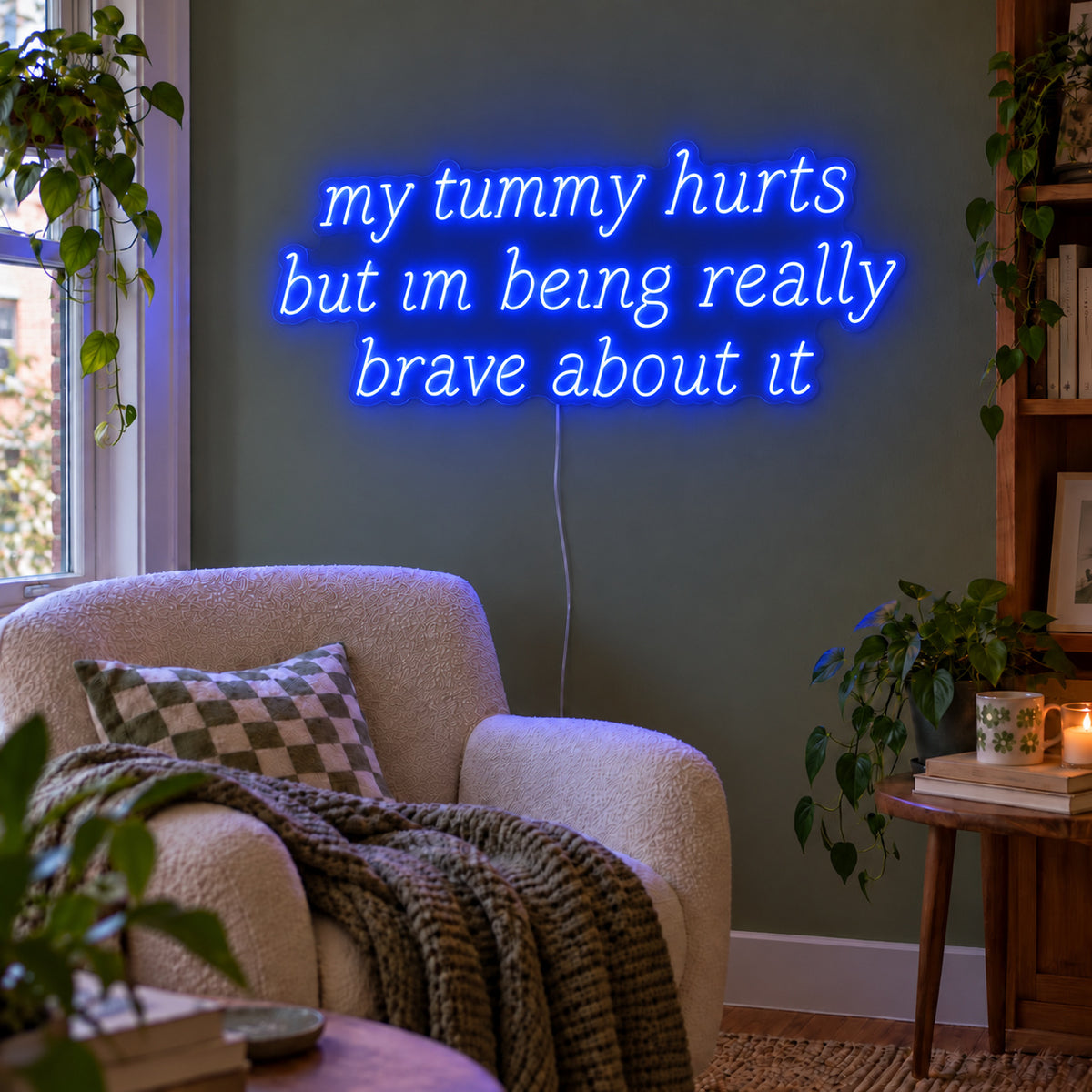 My Tummy Hurts But Im Being Really Brave About It Neon Sign glowing blue above a reading chair against a green wall.