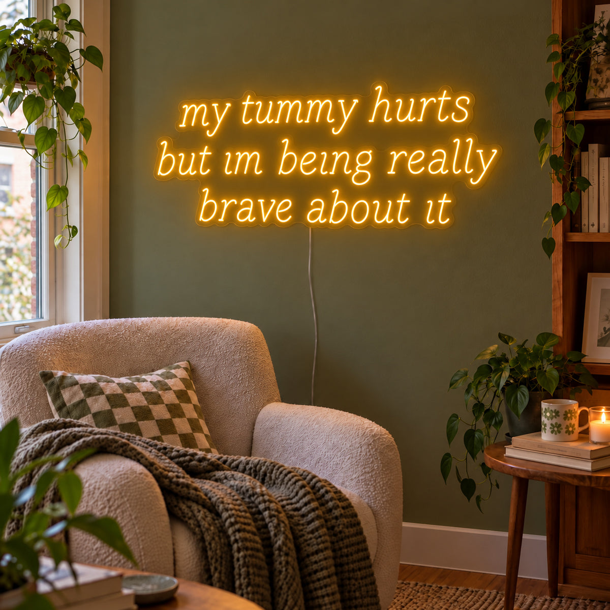 My Tummy Hurts But Im Being Really Brave About It Neon Sign glowing gold above a reading chair against a green wall.