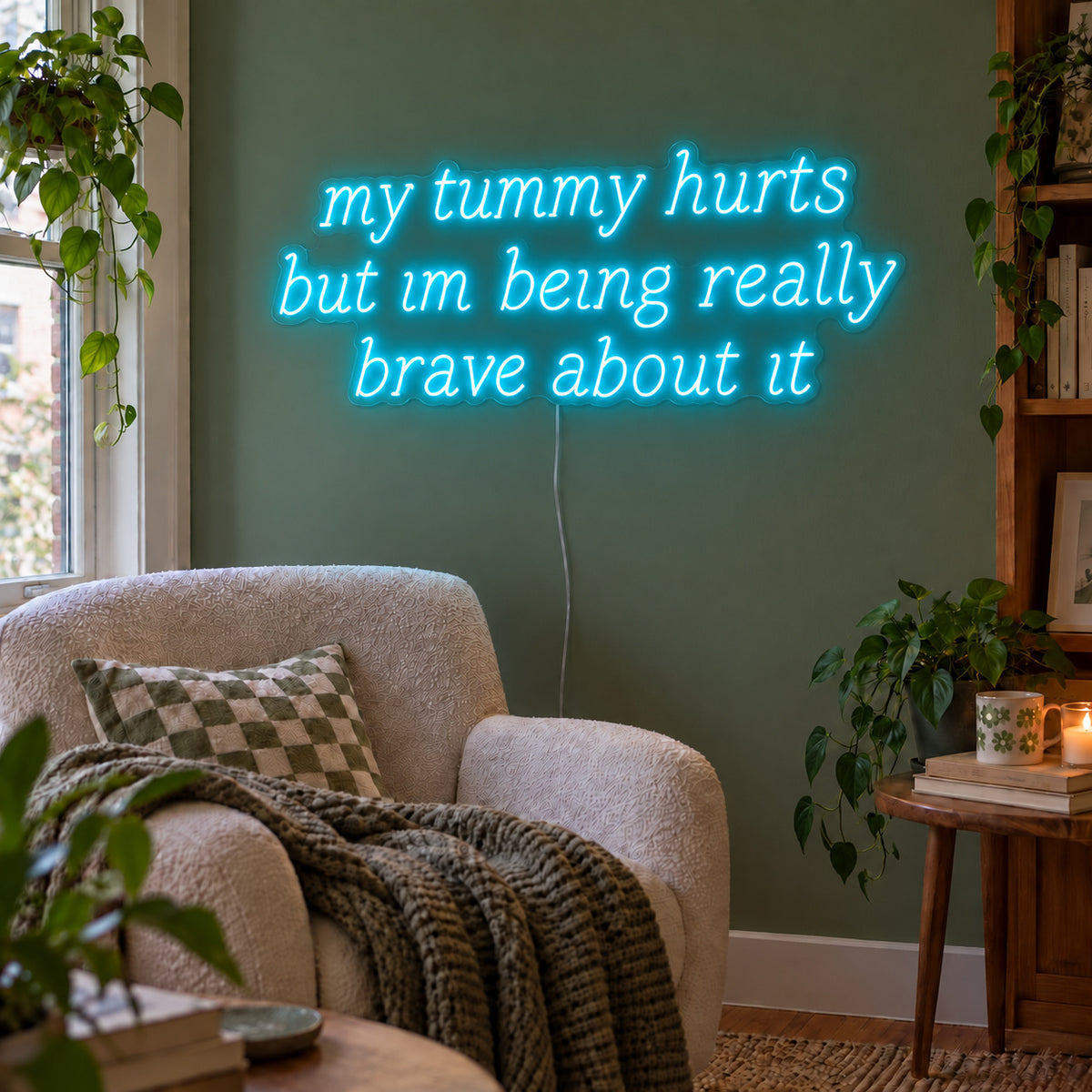 Ice Blue version of the My Tummy Hurts But Im Being Really Brave About It Neon Sign, shown above a reading chair against a green wall.