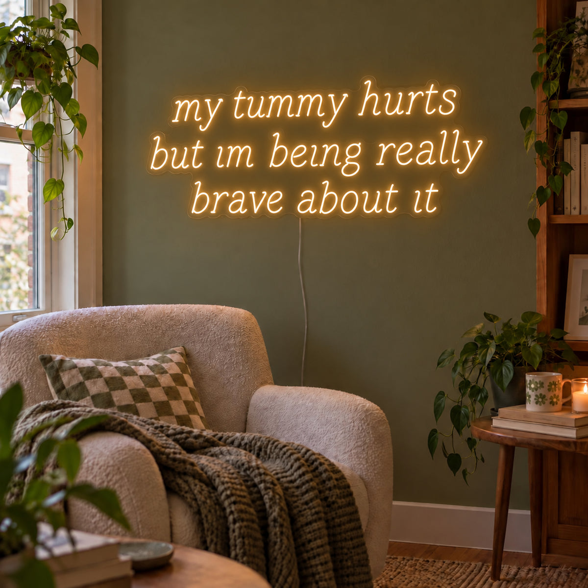 My Tummy Hurts But Im Being Really Brave About It Neon Sign in warm white above a reading chair against a green wall.
