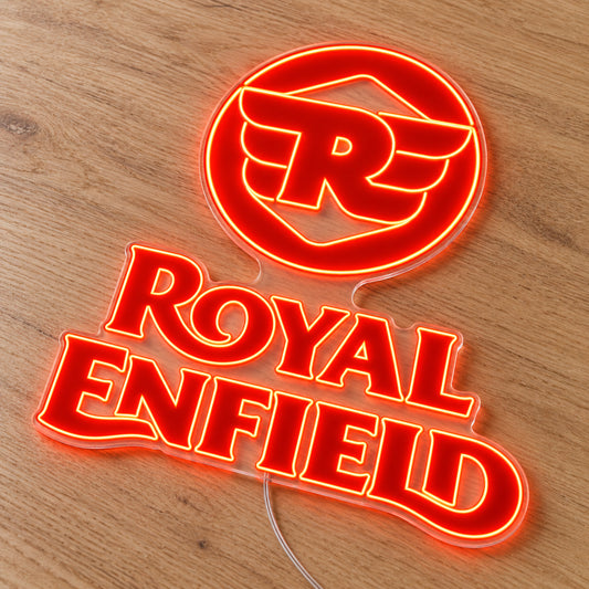 Royal Enfield neon sign with red-orange winged R logo and lettering on a single clear acrylic backboard, angled view on a light wooden surface.