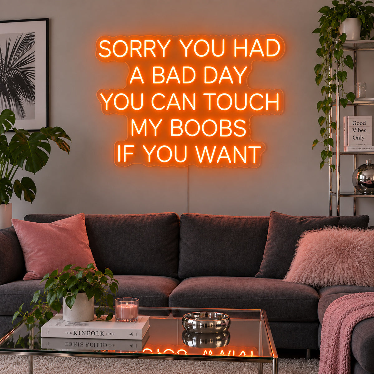 Orange version of the Sorry You Had A Bad Day You Can Touch My Boobs If You Want Neon Sign, shown above a grey sofa with pink cushions.