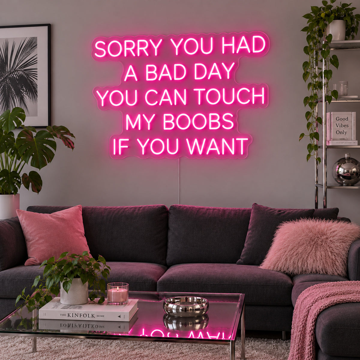 Rose Red version of the Sorry You Had A Bad Day You Can Touch My Boobs If You Want Neon Sign, shown above a grey sofa with pink cushions.