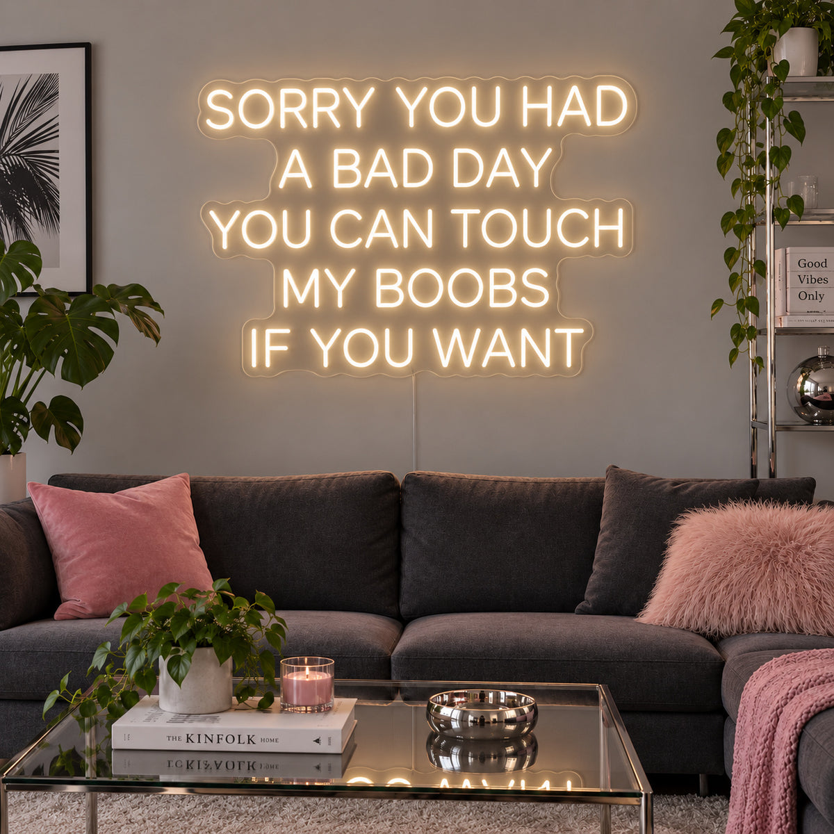 Warm White version of the Sorry You Had A Bad Day You Can Touch My Boobs If You Want Neon Sign, shown above a grey sofa with pink cushions.